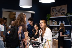 A Meantime cashier smiles at a customer who has their back turned to the camera. Two other Meantime employees can be seen in the background.