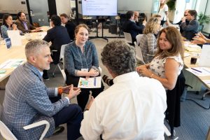 A group of four people sit in a circle with more people in the background.
